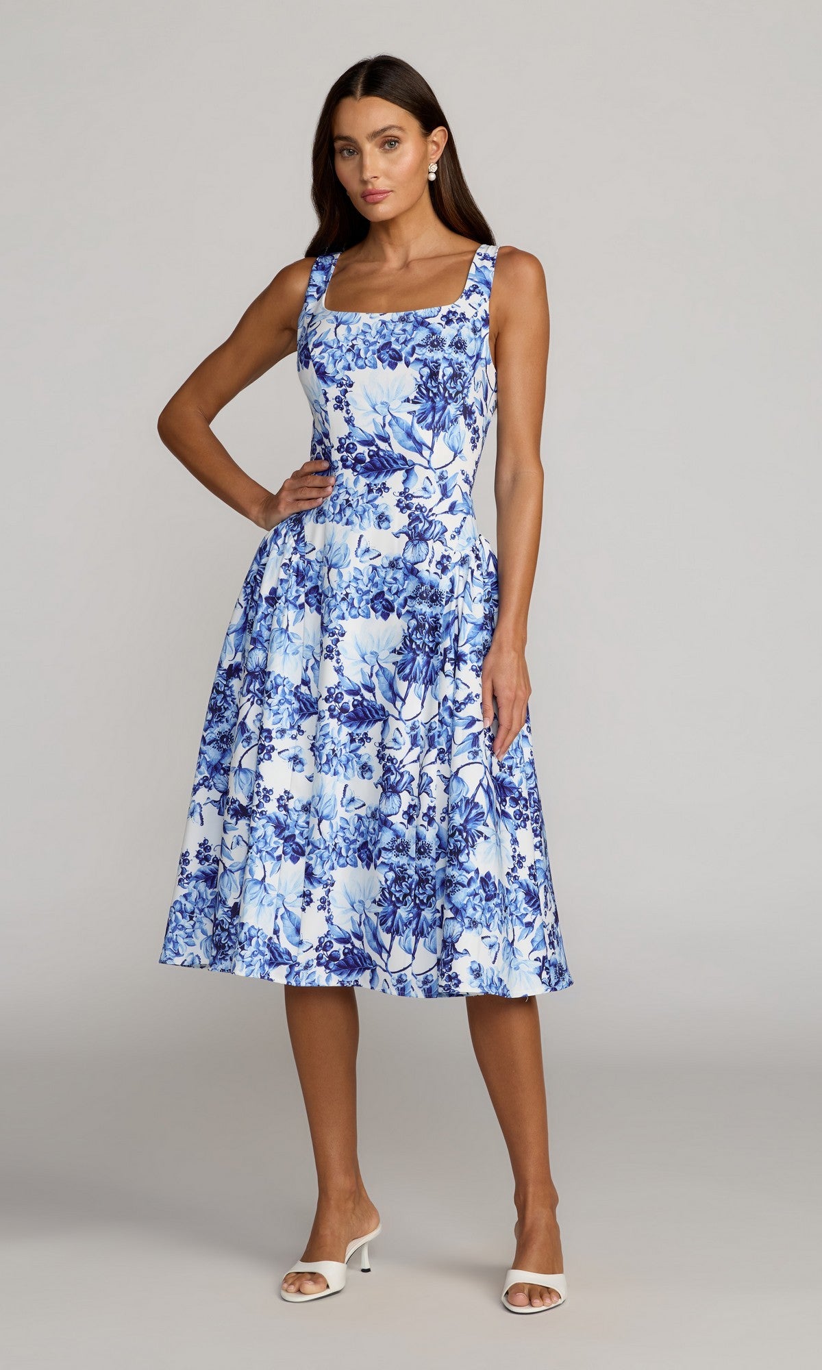 A woman wearing the Mac Duggal 37155 Floral Midi Wedding Guest Dress in Blue Multi with a sleeveless square neckline and white open-toe heels, standing against a plain light background.